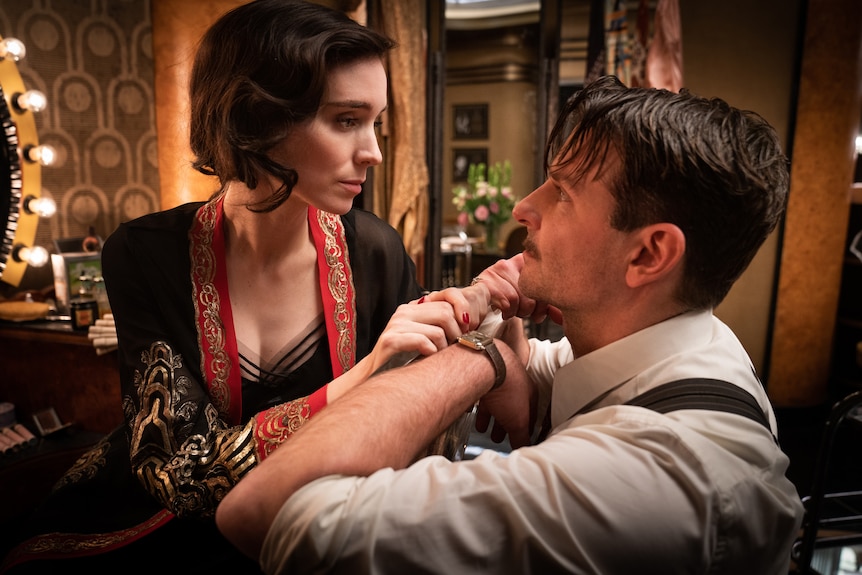 A 30-something brunette woman looks concerns as she stares into the eyes a determined 40-something man, each dressed in 30s garb
