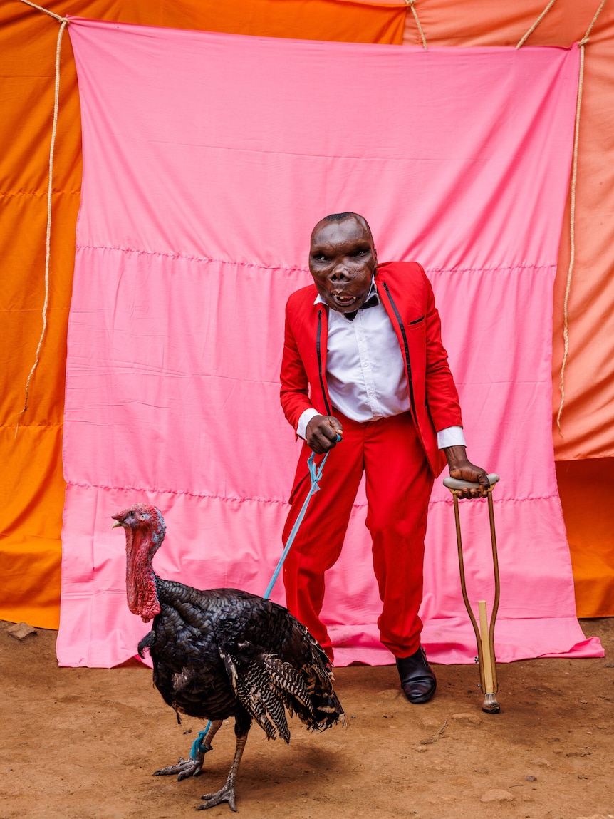 A short man stands with a crutch, holding a turkey on a rope lead in front of bright pink and orange sheets.