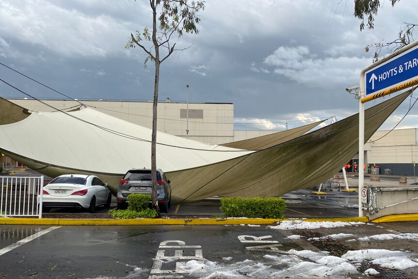 A large sunshade awning sags over two cars in a car park. 
