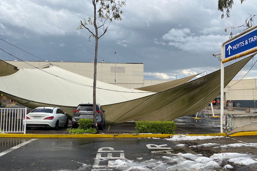 A large sunshade awning sags over two cars in a car park. 