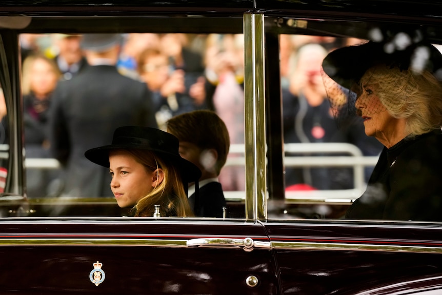 Princess Charlotte sits in a car, Prince George is in the backgroud and Camilla, the Queen Consort, behind. 