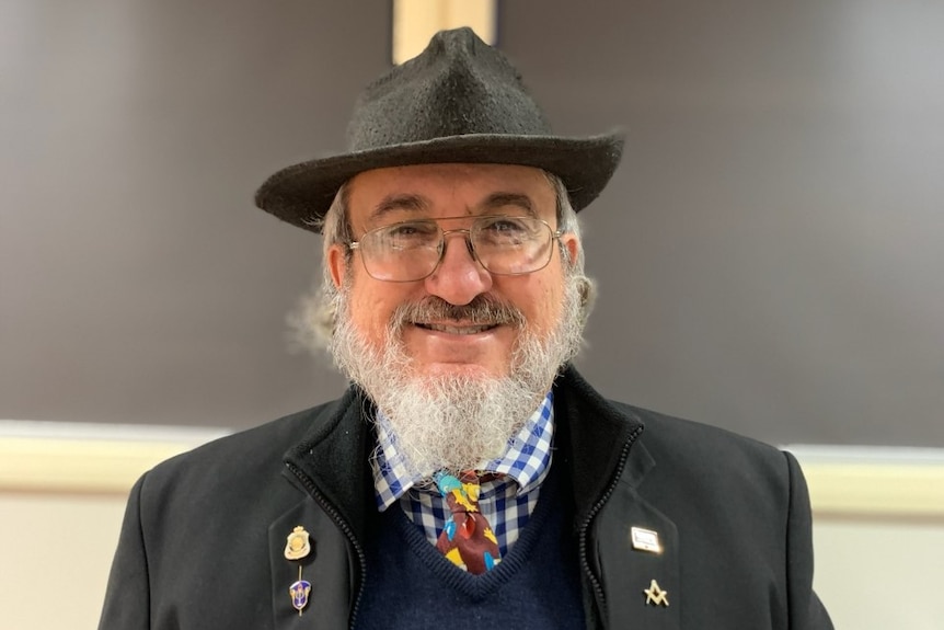 A man with glasses and a grey beard smiles. He is wearing a grey fedora, and a black jacket.