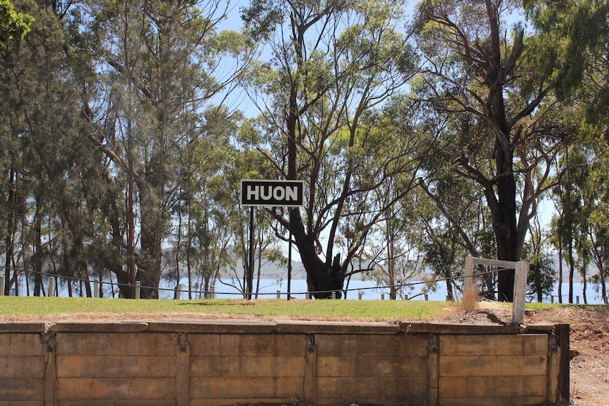 A black sign states Huon in white letters. It stands on what remains of the railway platform, backed by trees and Lake Hume.
