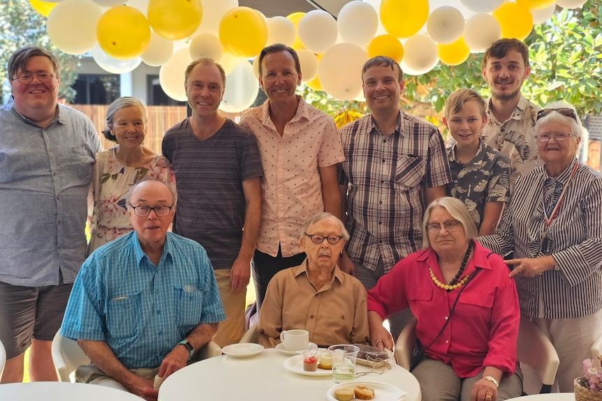 An elderly man flanked by family members with balloons in the background.