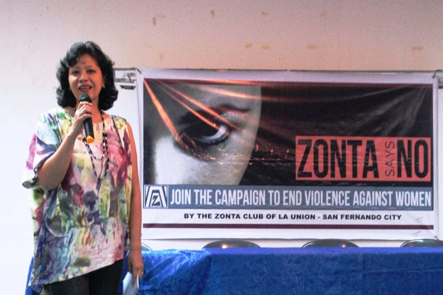 A woman holding a microphone in front of a sign which reads: Join the campaign to end violence against women