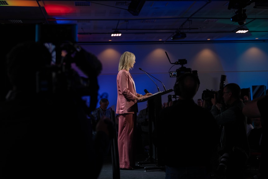 A blonde woman in a pink suit stands at a podium in a dark, crowded room.