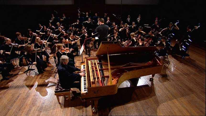 A man plays the piano, surrounded by a large orchestra