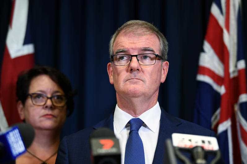 A man in a suit stands in front of microphones looking forlorn and a woman stands in the background.