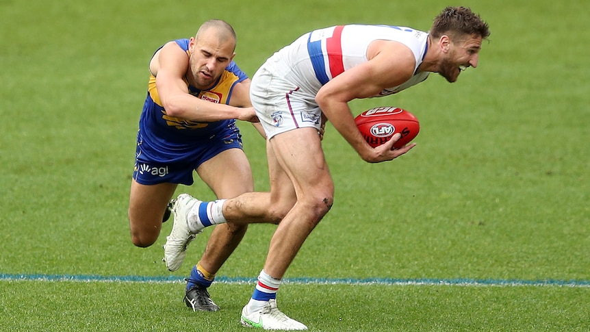 A Western Bulldogs AFL player holds the ball with his right hand as he is tackled by a West Coast opponent.