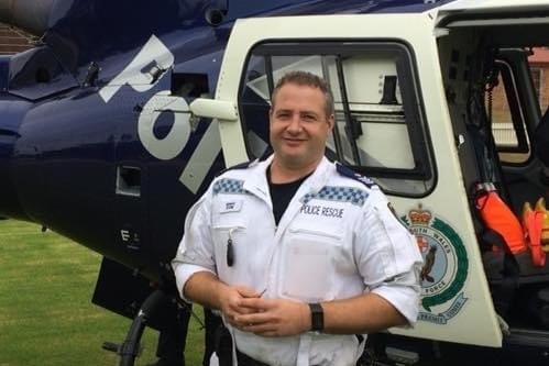 A man wearing a police rescue uniform smiles in front of a helicopter