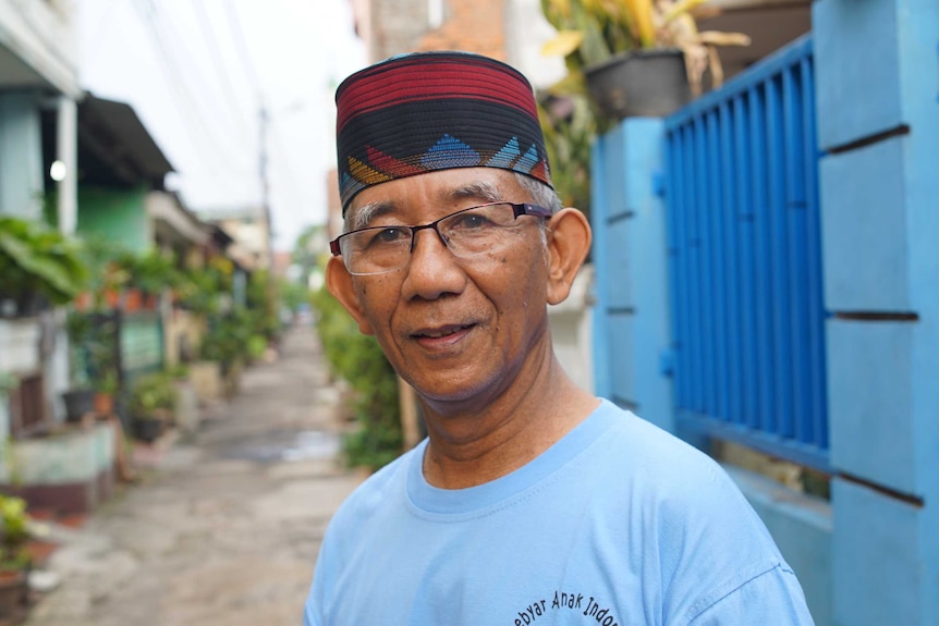 Andang Subaryono, the head of Perumnas Klender. He is standing in an alleyway wearing a red and black fez, glasses and blue tee.