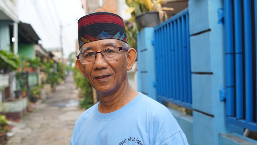 Andang Subaryono, the head of Perumnas Klender. He is standing in an alleyway wearing a red and black fez, glasses and blue tee.