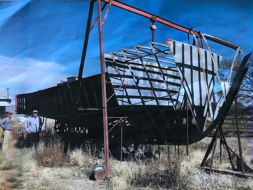 A large boat under construction sits within a metal frame on dry grass with two men standing nearby.