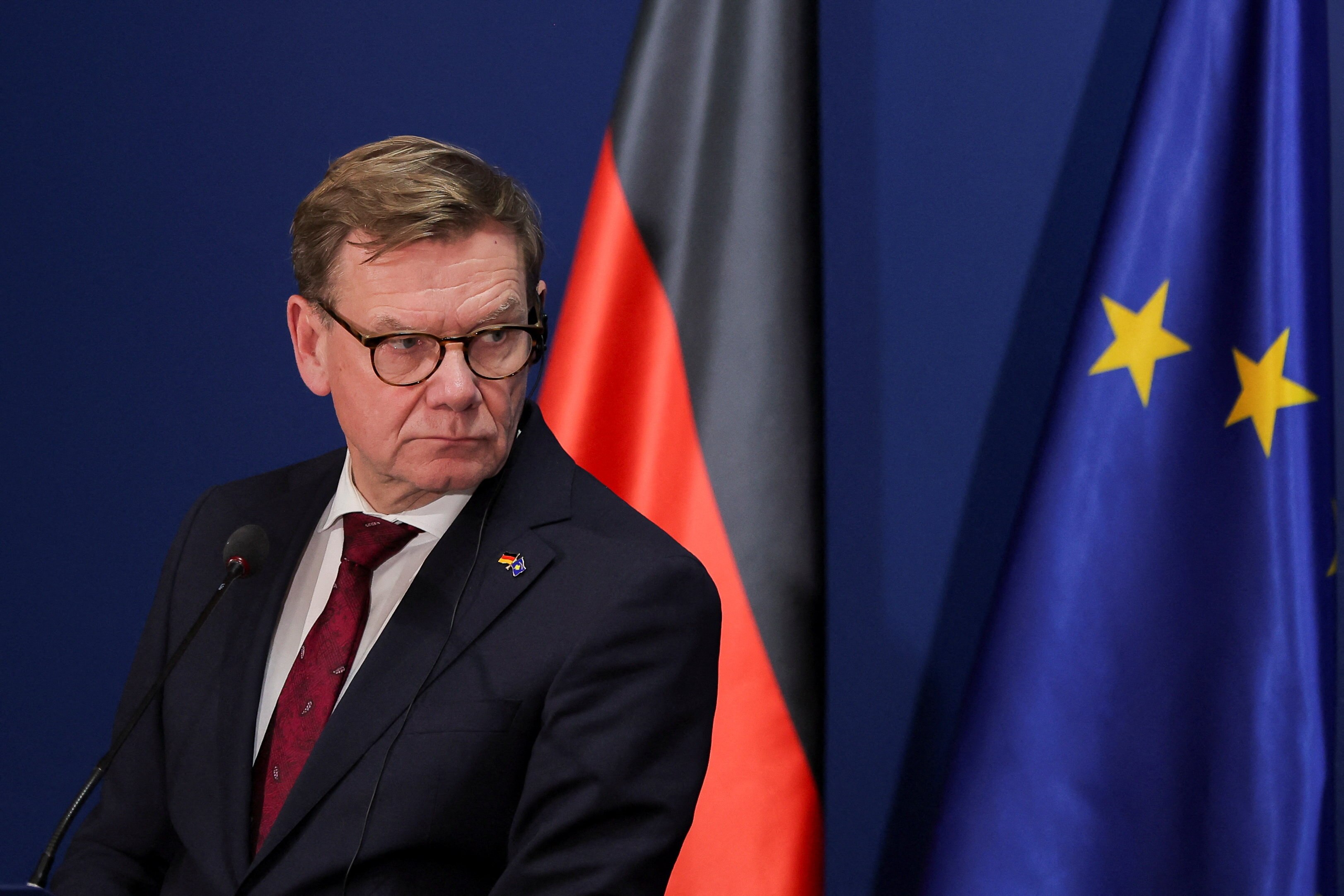 Un hombre de mediana edad con gafas se encuentra con expresión severa frente a una bandera alemana y una de la UE.