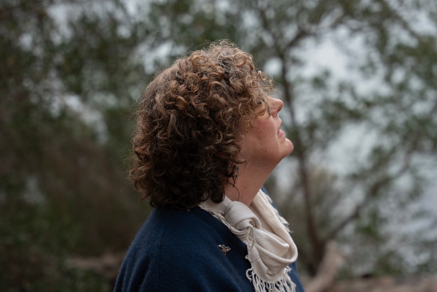 A woman with curly brown hair looks to the sky, dark bush and water blurred in background.