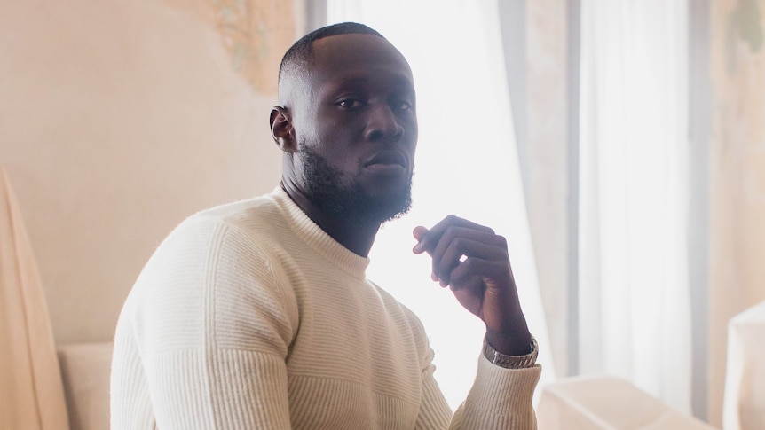 A 2022 press shot of Stormzy on a stool in a white crewneck sweater