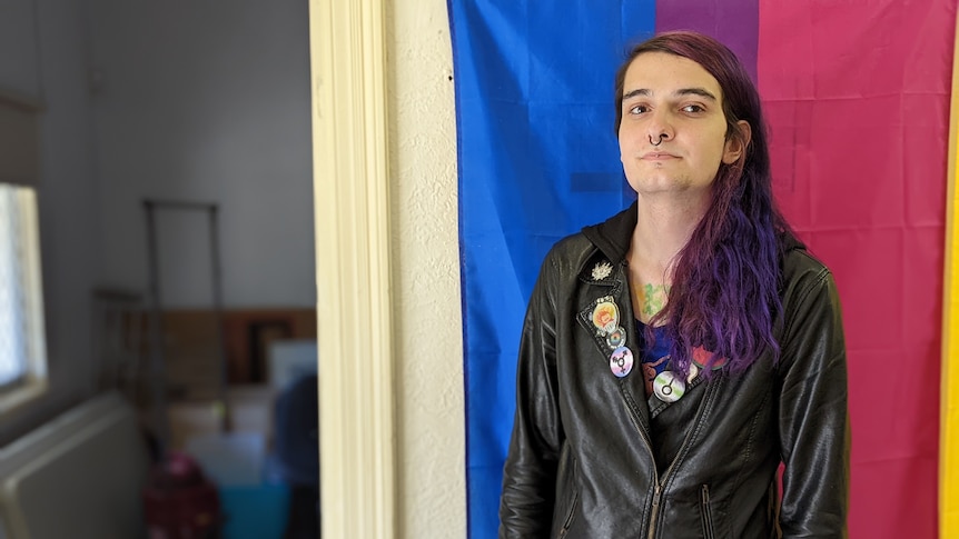 Opal has long purple hair and wears leather jacket with badges standing in front of colourful flag inside a house. 