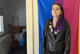Opal has long purple hair and wears leather jacket with badges standing in front of colourful flag inside a house. 