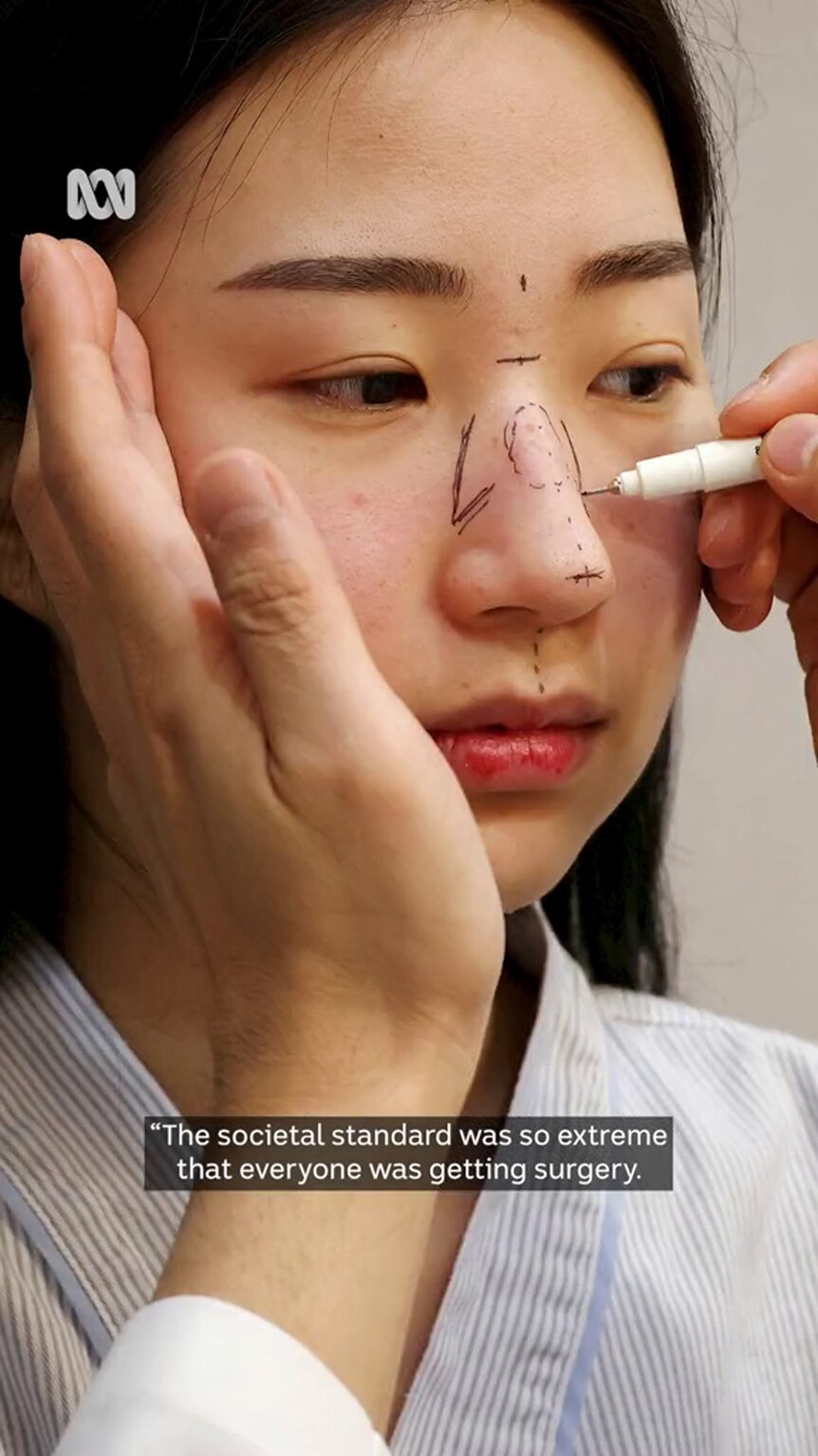 A young Asian woman has markings drawn on her face by hands with a skin tone that matches hers
