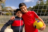 Brothers Zayn and Meeka hold soccer balls and embrace each other at a sports field.