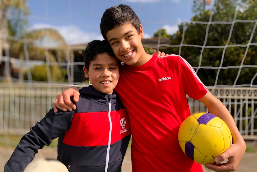 Brothers Zayn and Meeka hold soccer balls and embrace each other at a sports field.