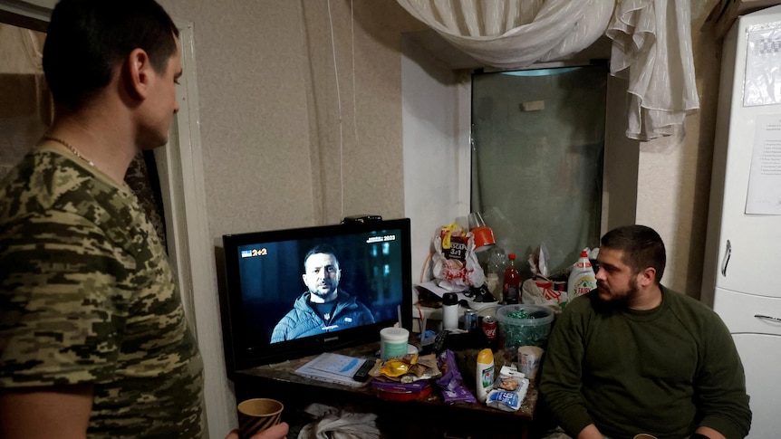 Two Ukrainian soldiers watch President Zelenskyy's new year's eve address to the nation on a small TV.