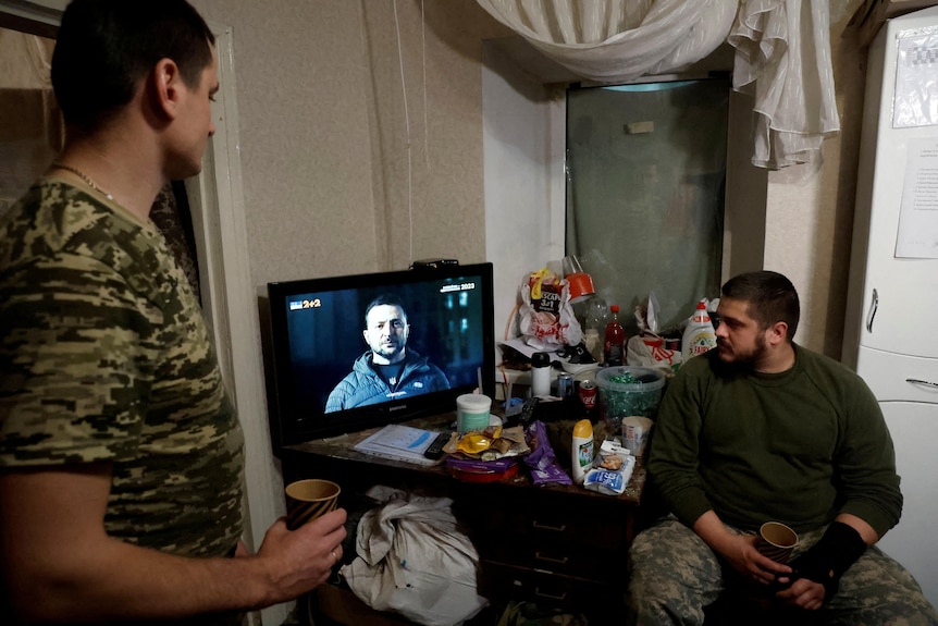 Two men in fatigues watch a television showing Vlodomyr Zelenskyy