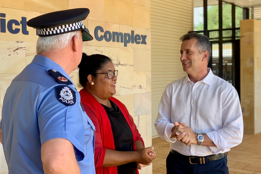 A woman with dark hair and a red shirt speaks with a uniformed police officer and a man in a white shirt