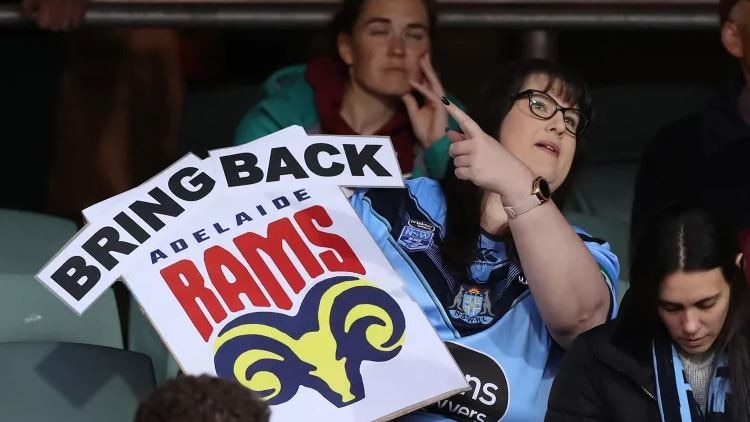 lady in spectacles and blue rugby league guernsey holding a sign saying 'Bring Back Adelaide Rams' 