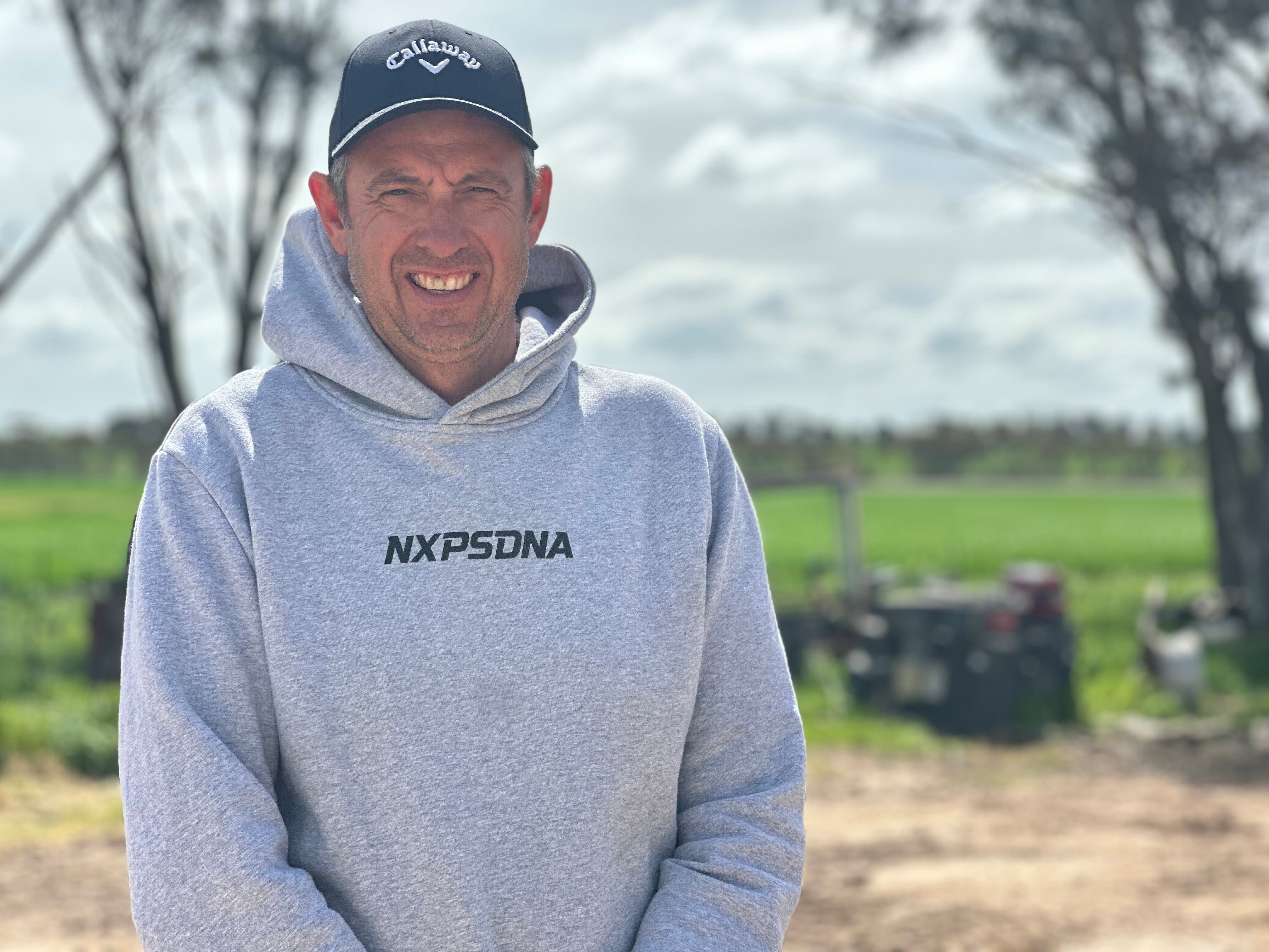 A man wearing a grey hoodie and blue hat stands in front of a green field.