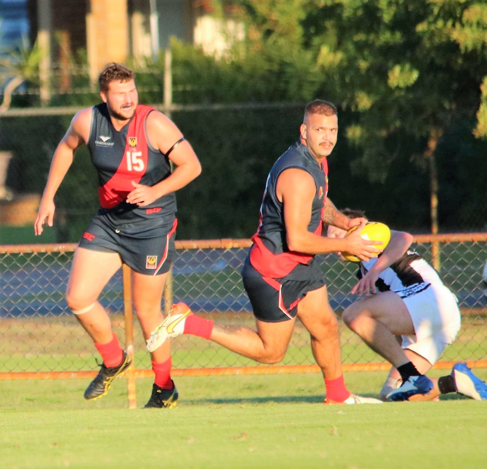 A footballer in black jumper and red sash looks downfield as he runs with the a ball  