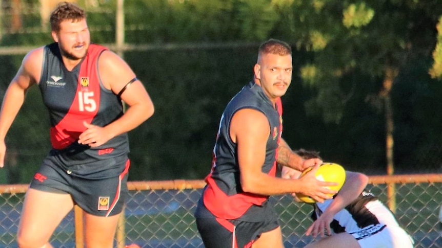 An Aboriginal footy player holds onto a ball planning his next move while a teammate approaches. 