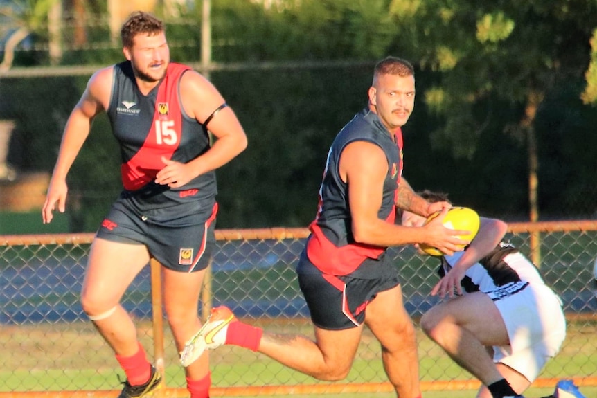 A footballer in black jumper and red sash looks downfield as he runs with the a ball  