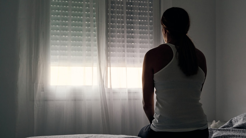 Rear view of an unrecognizable abused woman sitting on her bed looking out the window. Concept of gender violence,