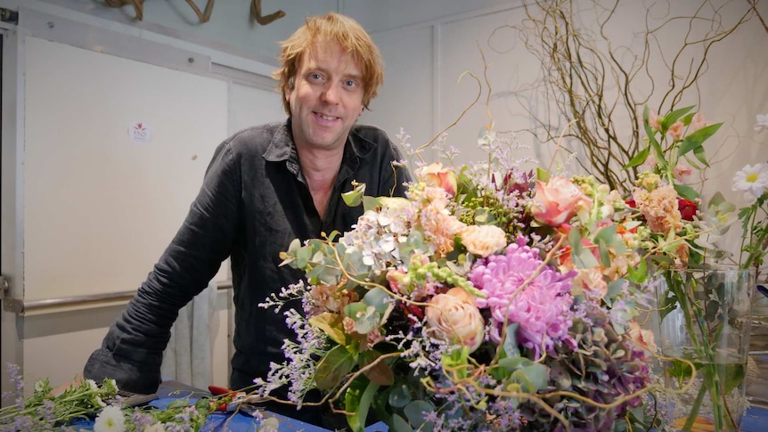 Man standing behind a table covered in flowers and big floral arrangement.