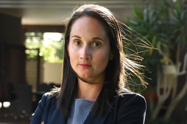 Woman with straight brown hair stands unsmiling with arms crossed.