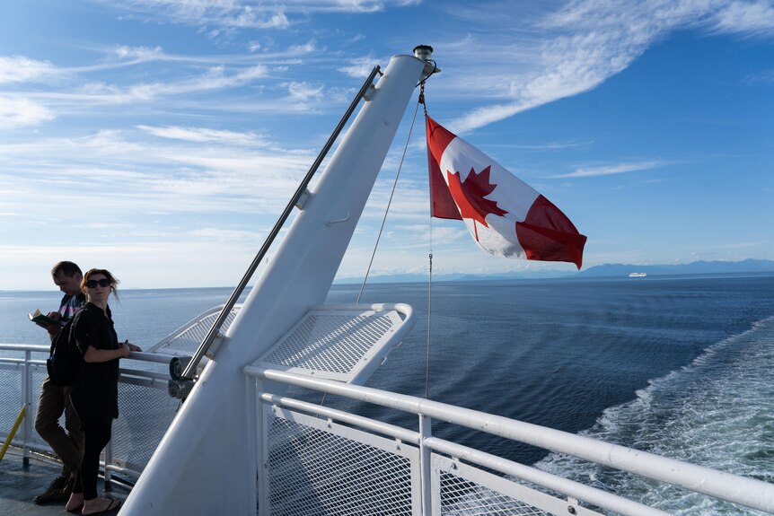 A Canadian flag hands off the end of a ferry against a blue sky and water. Two tourists stand nearby.