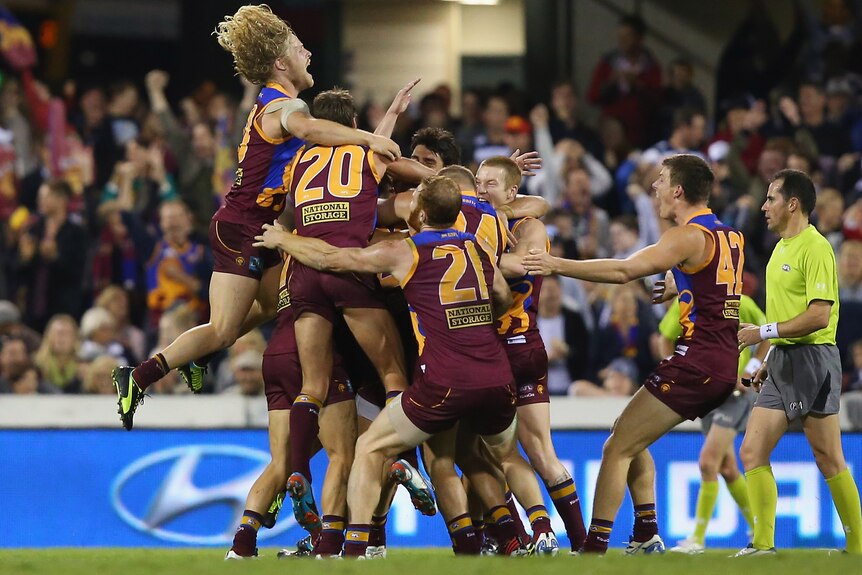 A group of Brisbane Lions AFL players rush into a ceklebratory huddle after a game, with some players leaping on from the side.