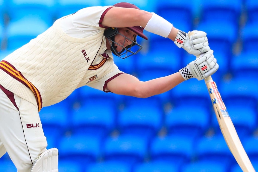 Cricketer Matthew Renshaw grimaces as he leans over with his bat out in front. He wears a red helmet to match his uniform trim.