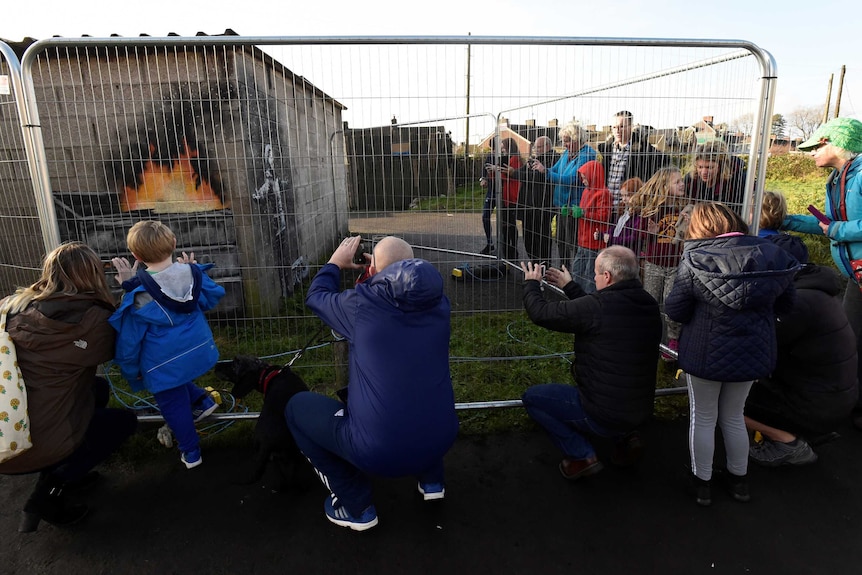 Crowds gather behind a steel fence that is protecting a Banksy artwork on a garage wall