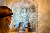 A woman wearing a headlamp works to restore an old picture on a wall