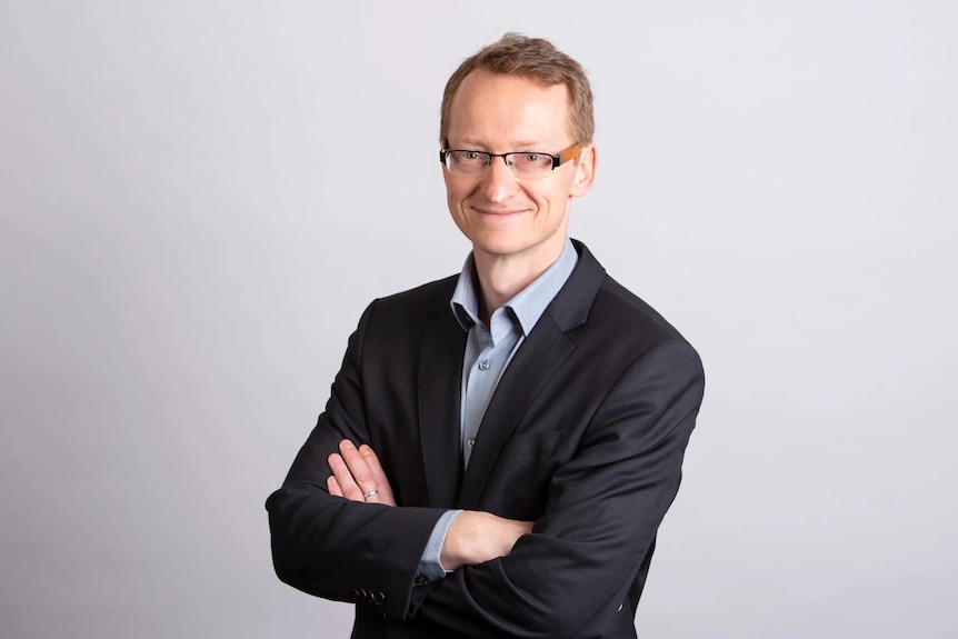 A middle-aged white man with short hair and glasses stands, wearing a business suit and smiling