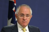Malcolm Turnbull shrugs and frowns while standing at a lectern. There are two Australian flags in the background