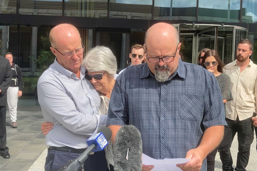 Mark looks at a piece of paper, a sad looking elderly woman standing behind him, hugging another man.