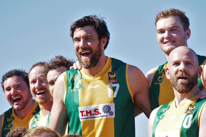 Man in green and gold uniform flanked by team mates laughs at camera