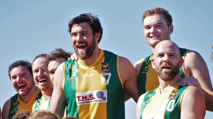 Man in green and gold uniform flanked by team mates laughs at camera