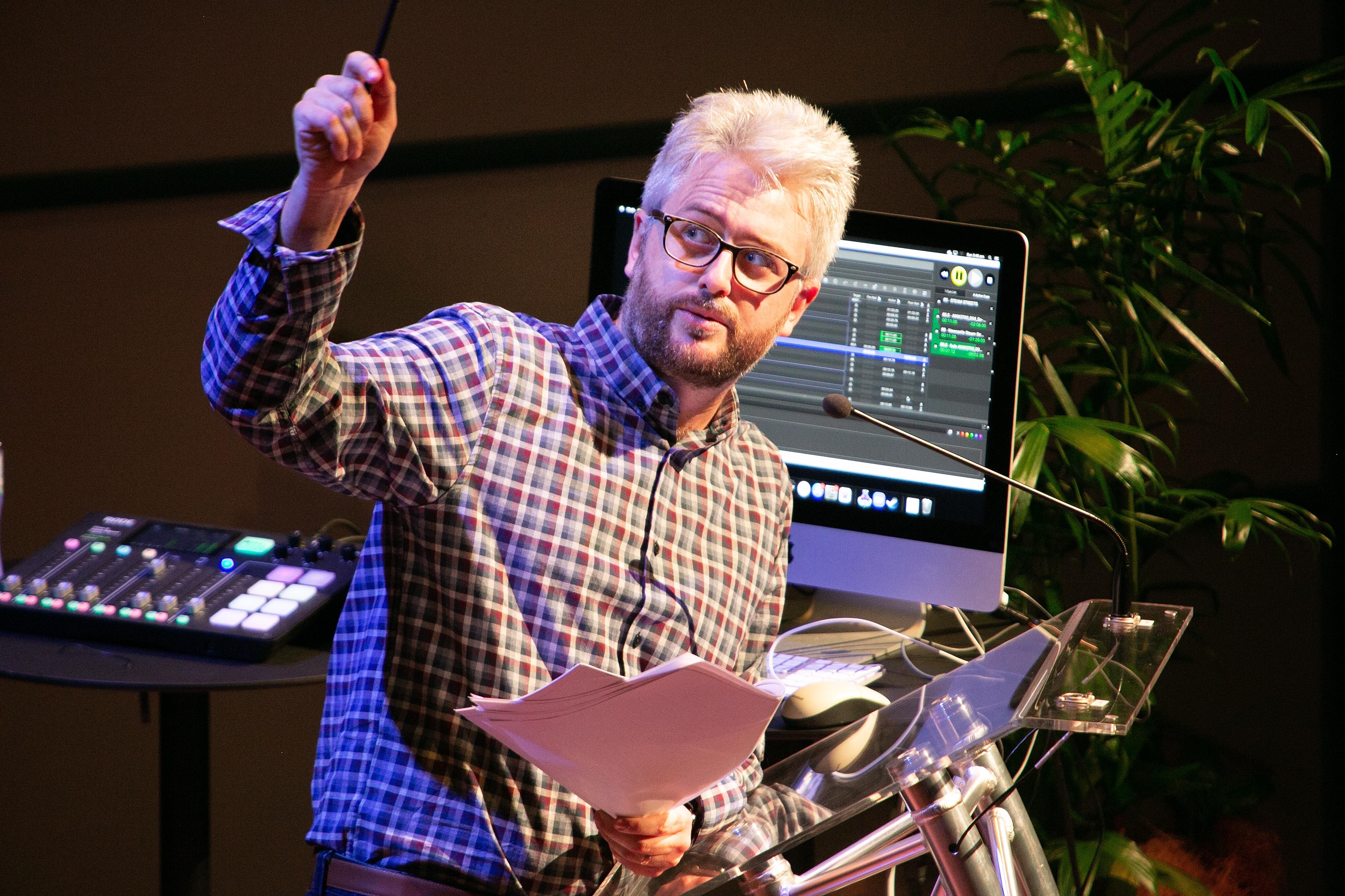 Man standing behind a microphone with computer screen behind him, looking off to the left of shot and pointing.