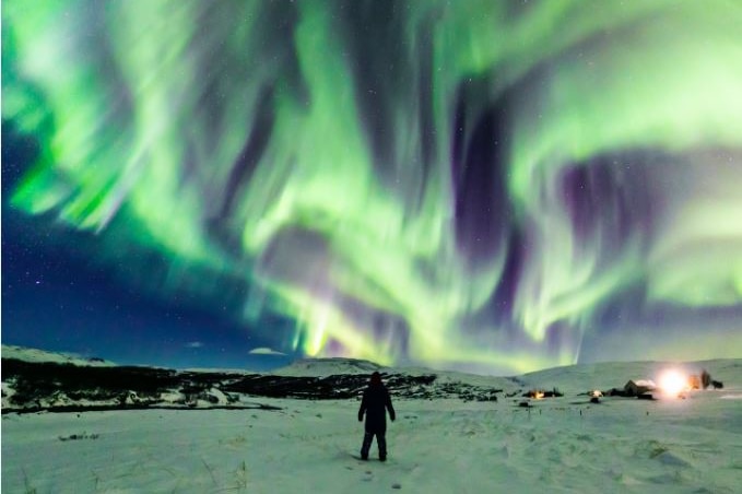 A figure stands in the snow, while green and purple lights streak the night sky, forming the shape of a dragon's head.