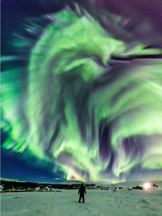 A figure stands in the snow, while green and purple lights streak the night sky, forming the shape of a dragon's head.
