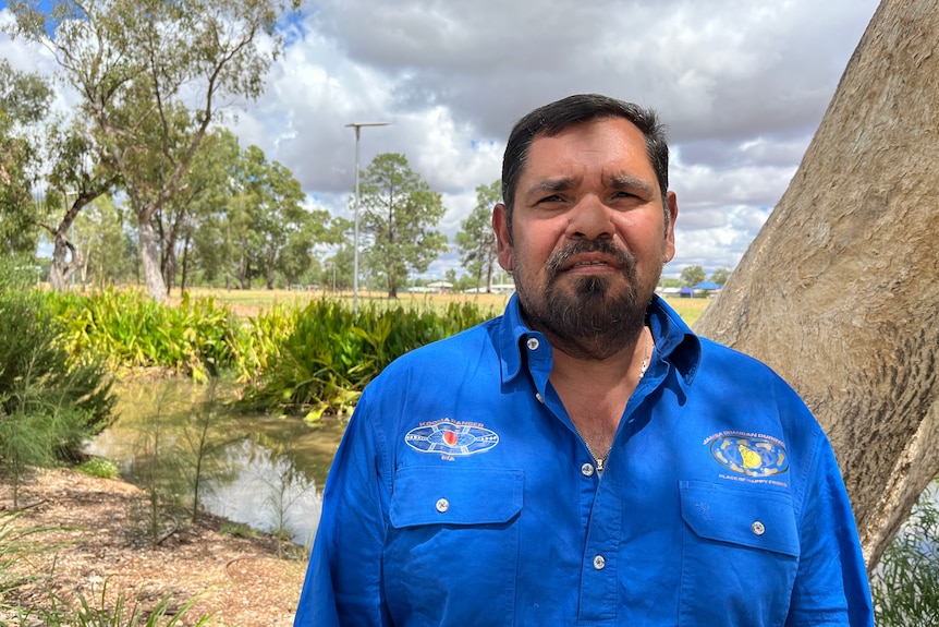 Ross Mitchell wears a blue work shirt and looks at the camera, in front of a water way and gum trees. 
